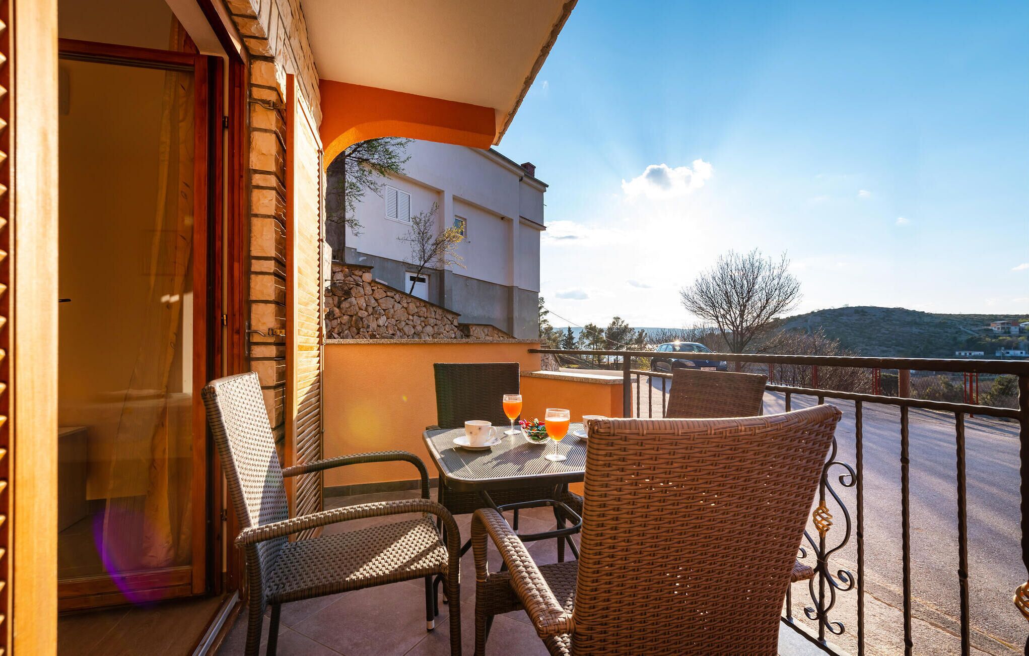 Un balcon avec des fauteuils en osier, une table avec des boissons et une vue sur un immeuble et un paysage.