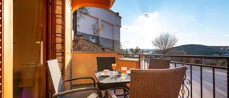 Ein Balkon mit Korbstühlen, einem Tisch mit Getränken und Blick auf ein Gebäude und die Landschaft.