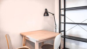 Una pequeña mesa de madera con dos sillas, una lámpara de escritorio negra y una estantería metálica negra junto a una pared blanca.