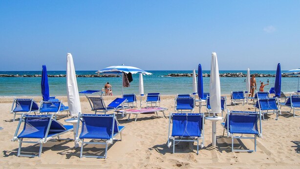 Une plage bordée de rangées de chaises longues bleues et de parasols blancs, un rivage rocheux et des eaux bleues et calmes.
