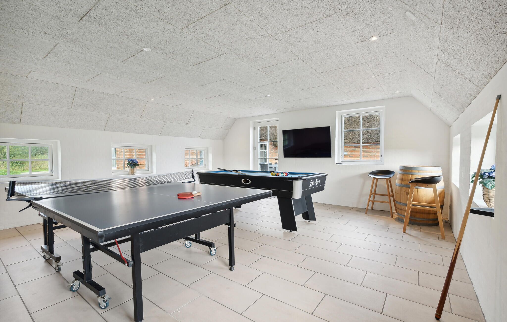 Amplia sala con mesa de ping pong, mesa de billar y mesa de futbolín. Hay ventanas con vistas a la vegetación, un televisor montado en la pared y un barril de madera.