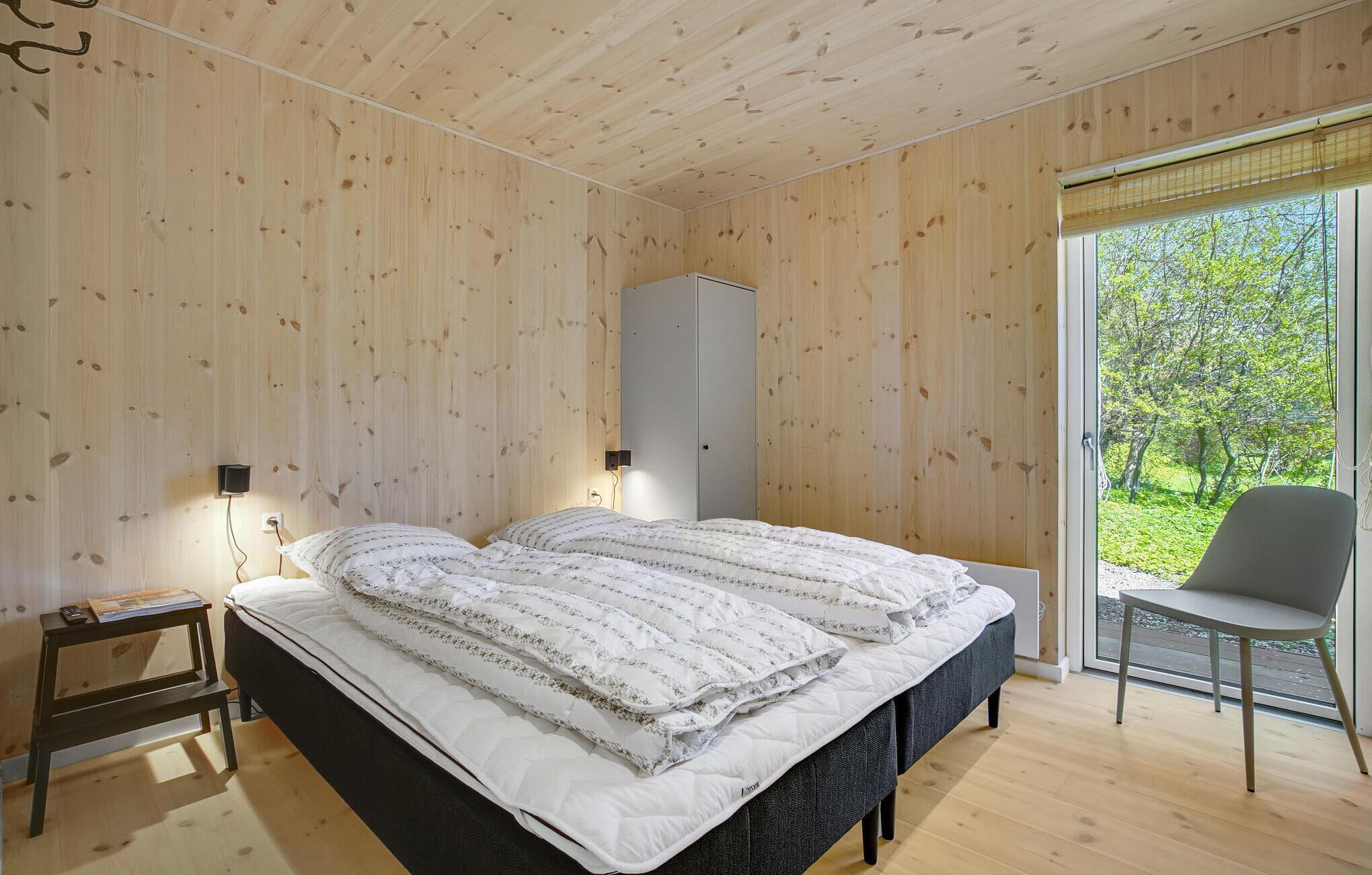 A bedroom with a bed, bedside tables, a wardrobe, and a view of greenery through a large window.