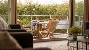 A balcony with a round wooden table and chairs, a glass of water, and a view of greenery.