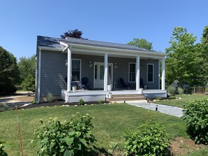 A single-story house with a covered porch, a front door, and a small garden area with plants and a lawn. - Custom cottage close to everything-Enjoy long walks on beach without the crowds! (Westerly)