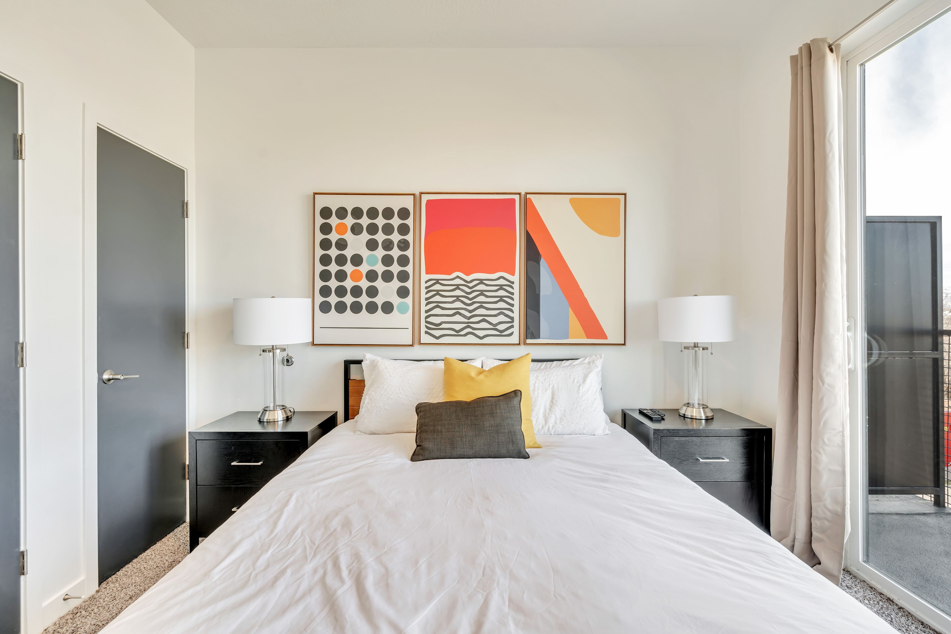 A hotel room with a large bed, two bedside tables with lamps, a window with curtains, and three framed artworks on the wall.