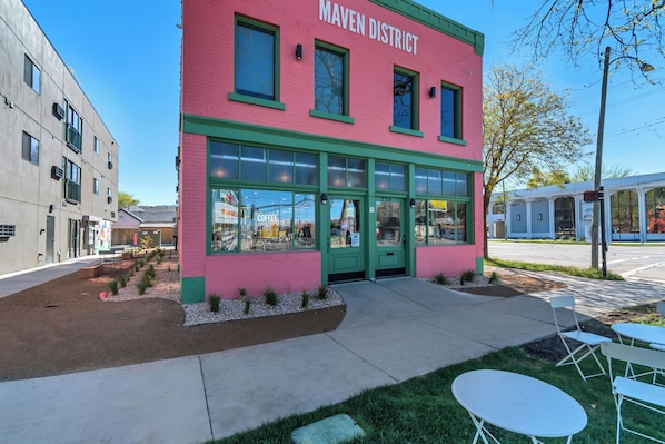 A building with a pink facade and green trim, labeled 'MAVEN DISTRICT,' featuring a storefront with large windows and a sidewalk in front.