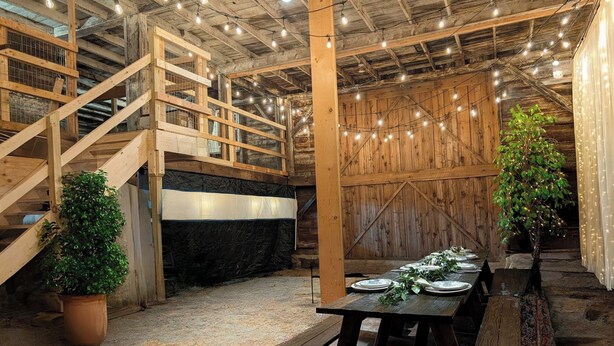 Un interior rústico con una escalera de madera, una mesa de comedor dispuesta con platos y luces colgantes en el techo.