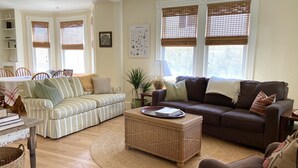 A living room with a striped sofa, a brown sofa, and a wicker coffee table. There are windows with blinds, a lamp, and a framed botanical print on the wall.