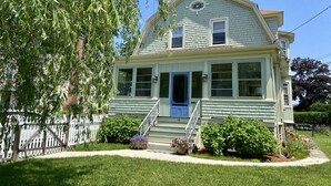 A two-story house with a front porch, a blue door, and a small front yard with a white picket fence and potted plants.