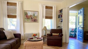 A living room with a brown sofa, a wicker coffee table with a vase of flowers, a brown armchair, a bookshelf, and a door leading outside.