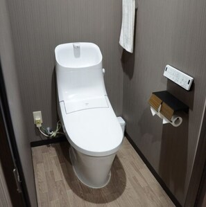 Une salle de bain compacte et moderne, avec des toilettes blanches, une petite étagère sur laquelle repose une boîte de mouchoirs et une prise électrique fixée au mur.