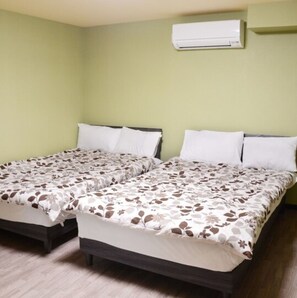 Une chambre avec deux lits, chacun recouvert d’une couvre-lit à motifs floraux, agrémenté d’oreillers blancs et doté d’une tête de lit en bois foncé. Un appareil de climatisation est fixé au mur, au-dessus des lits.