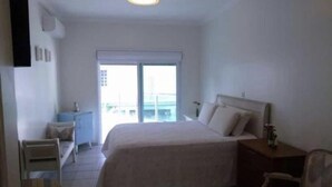 A hotel room with a large bed, a bedside table, a chair, a desk, and a window with a view of buildings. - Beautiful house in Peruibe (SP)