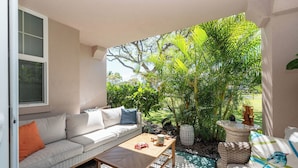 A cozy patio area with a white sofa, a wooden table, and a wicker chair. - 💎Luxury Home Away From Home! 🏝Beach🌞Sunshine🏌️‍♂️Golfing  (Waikoloa)