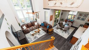 A spacious living area with a brown leather sofa, a coffee table, and a dining table with chairs. There is a staircase with a yellow railing leading to an upper floor. - 💎Luxury Home Away From Home! 🏝Beach🌞Sunshine🏌️‍♂️Golfing  (Waikoloa)