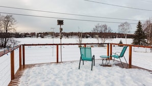 Eine Holzterrasse mit zwei Stühlen und einem Tisch, umgeben von einem Metallgeländer, mit Blick auf eine verschneite Landschaft mit Häusern und Bäumen im Hintergrund.