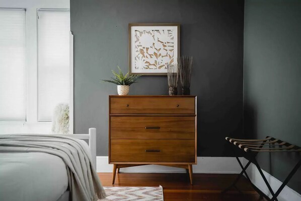 Une chambre à coucher avec un lit, une commode en bois, un tableau encadré, une plante en pot et une chaise.
