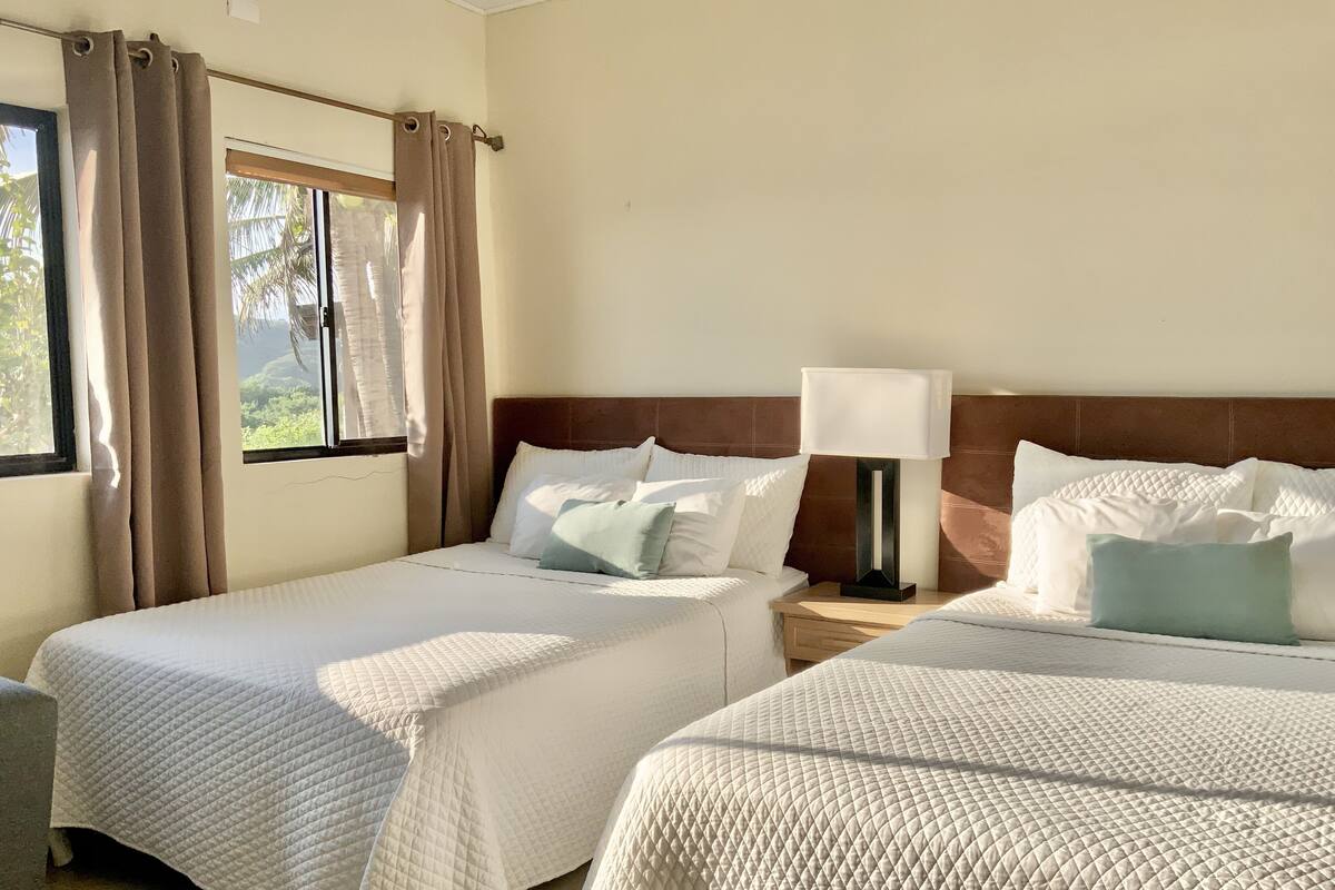 Dos camas individuales con ropa blanca, cabecero de madera, mesita de noche con lámpara y ventana con cortinas.