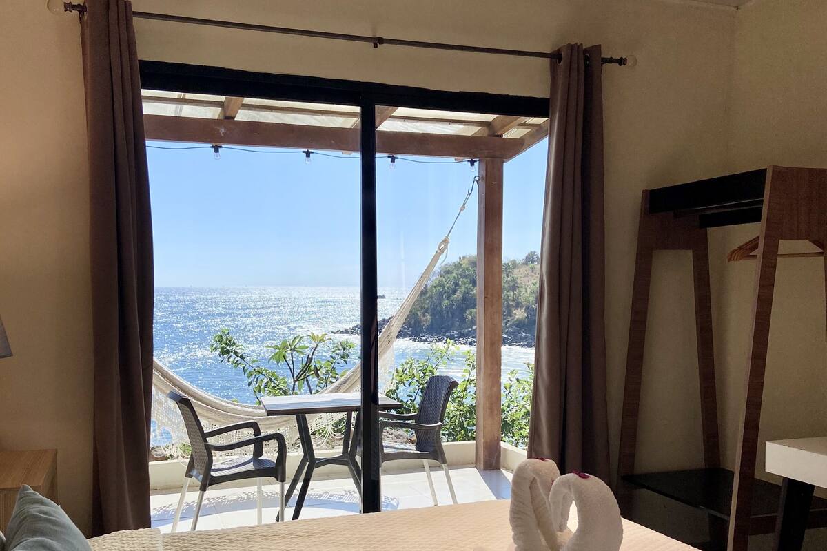 Una habitación de hotel con una cama, vista al océano y una hamaca afuera.