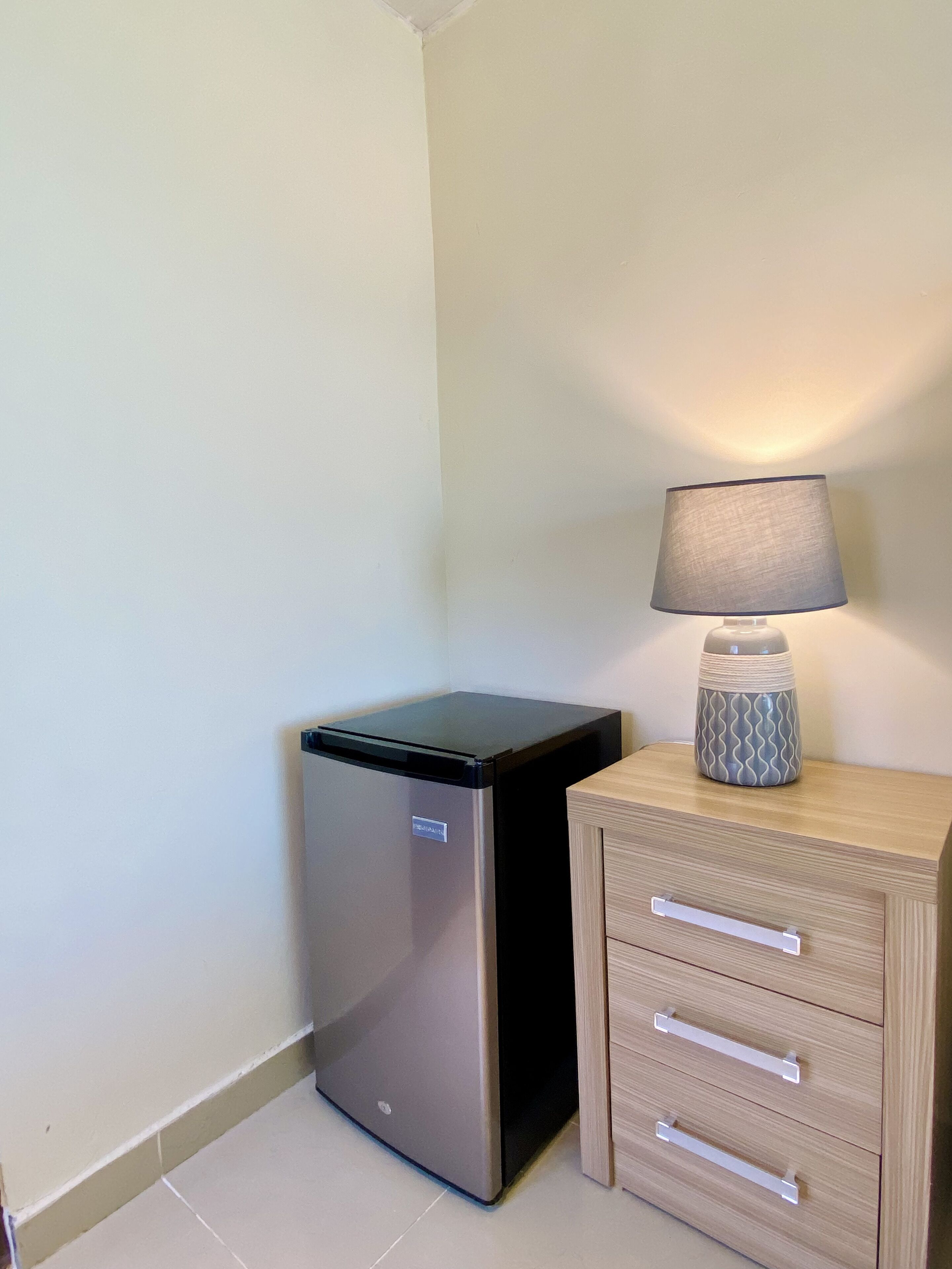 A compact refrigerator next to a wooden cabinet with drawers, a lamp on top, and a plain wall.