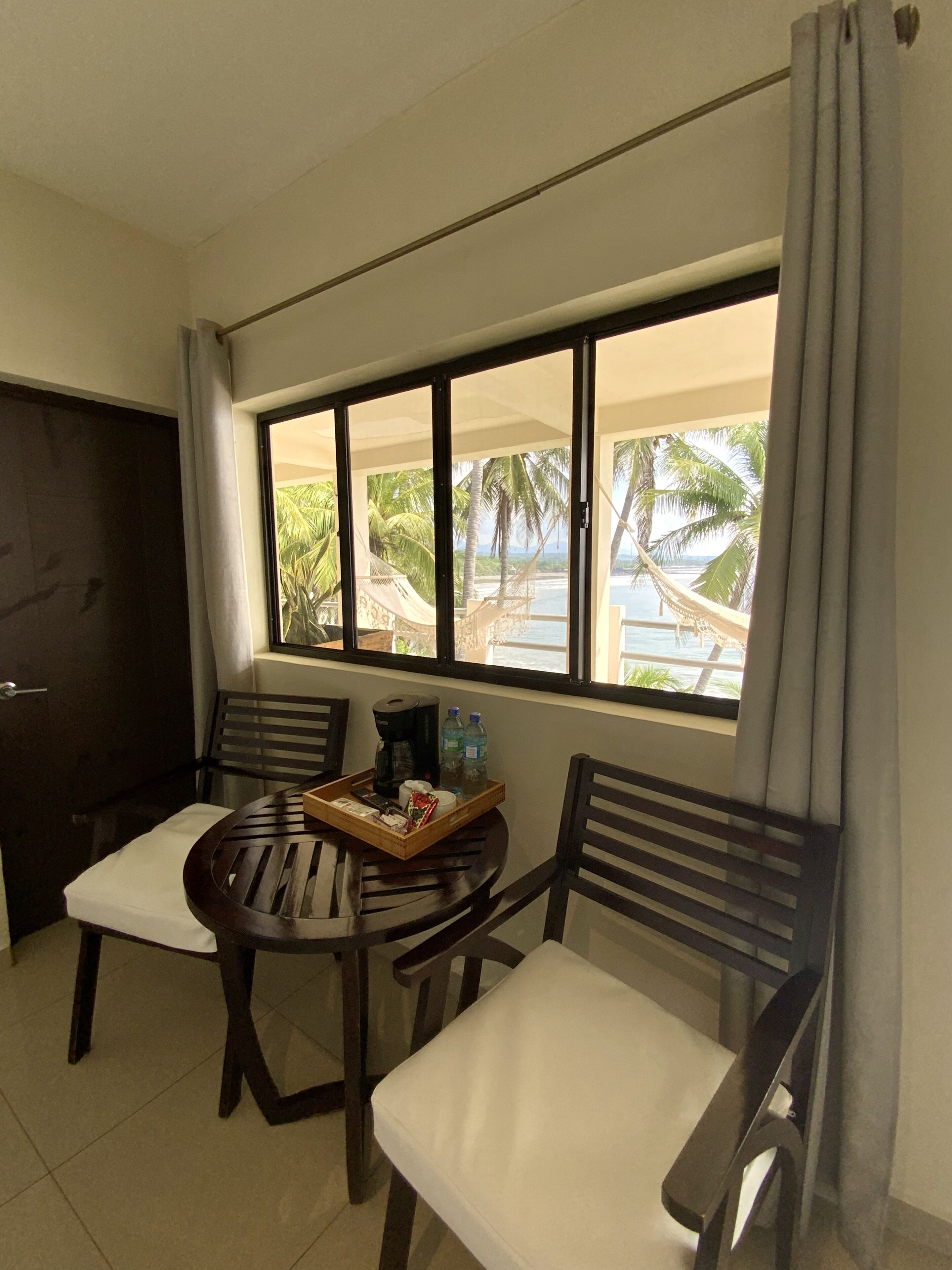 A room with a large window offering a view of a beach and palm trees. There is a round wooden table with chairs, a tray with a coffee maker, and a bottle of water.