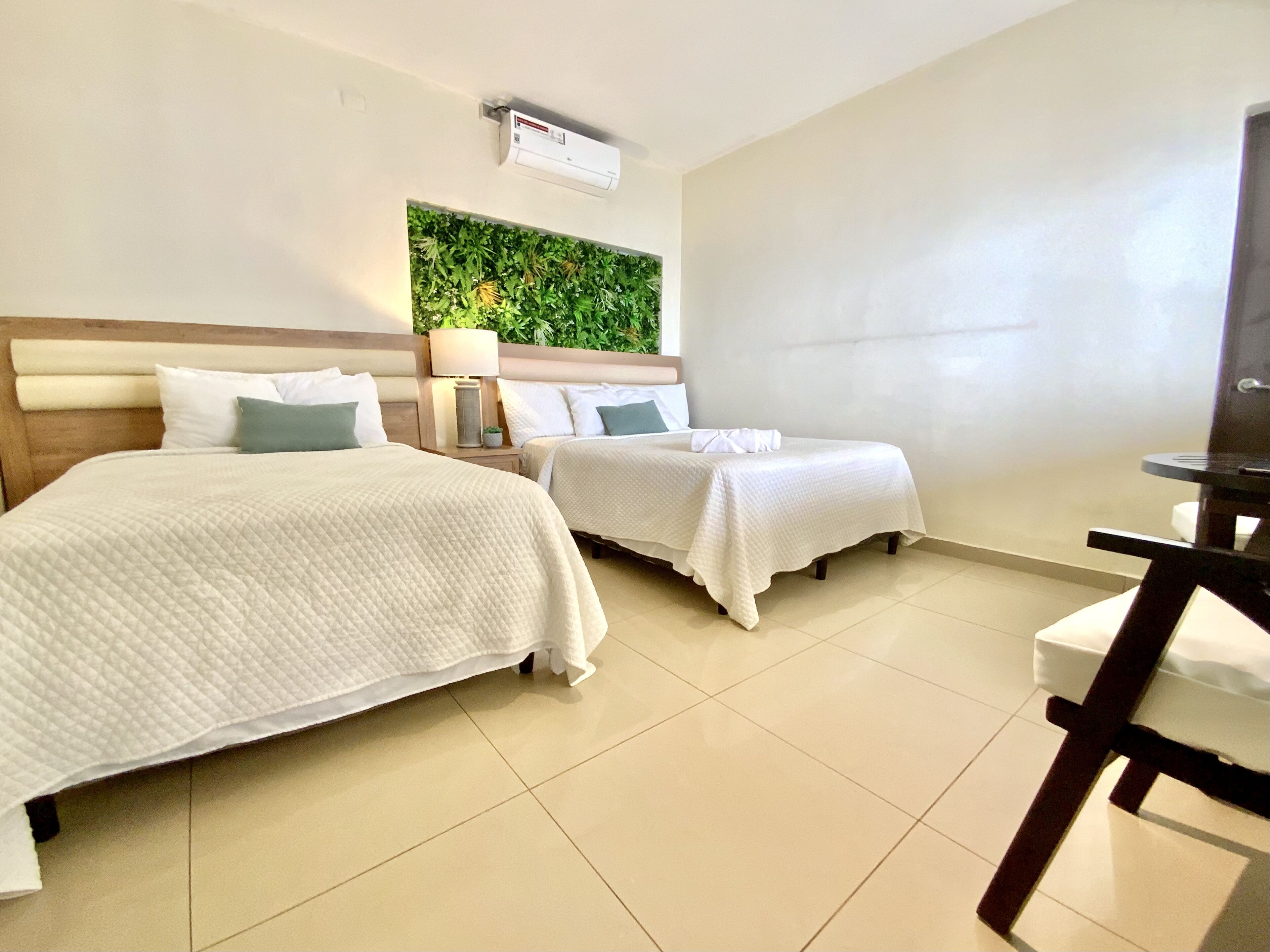 Une chambre d’hôtel avec deux lits, un mur vert orné de plantes, un climatiseur au plafond et une petite table avec une lampe.