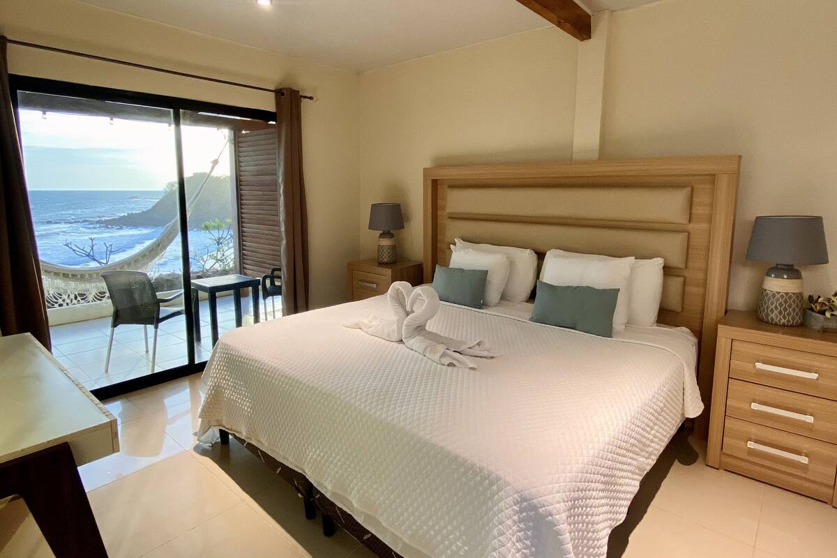 Un dormitorio espacioso con una cama grande, un cabecero de madera, dos mesitas de noche con lámparas y vistas al océano a través de un ventanal corredera.