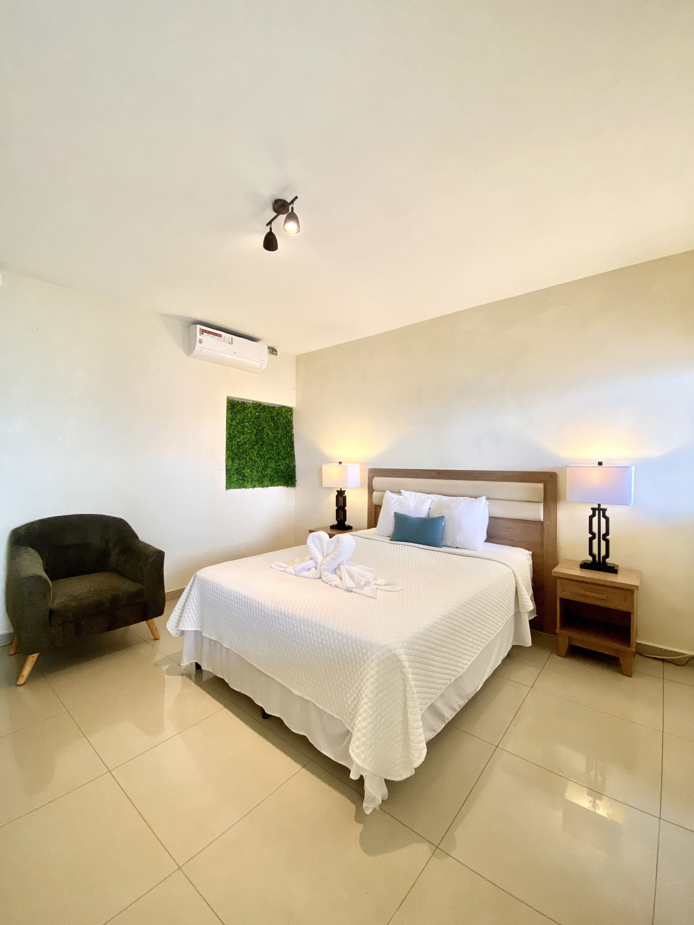 Quarto de hotel com cama, uma cadeira, duas mesas de cabeceira com luminárias, ar condicionado e um painel decorativo verde na parede.