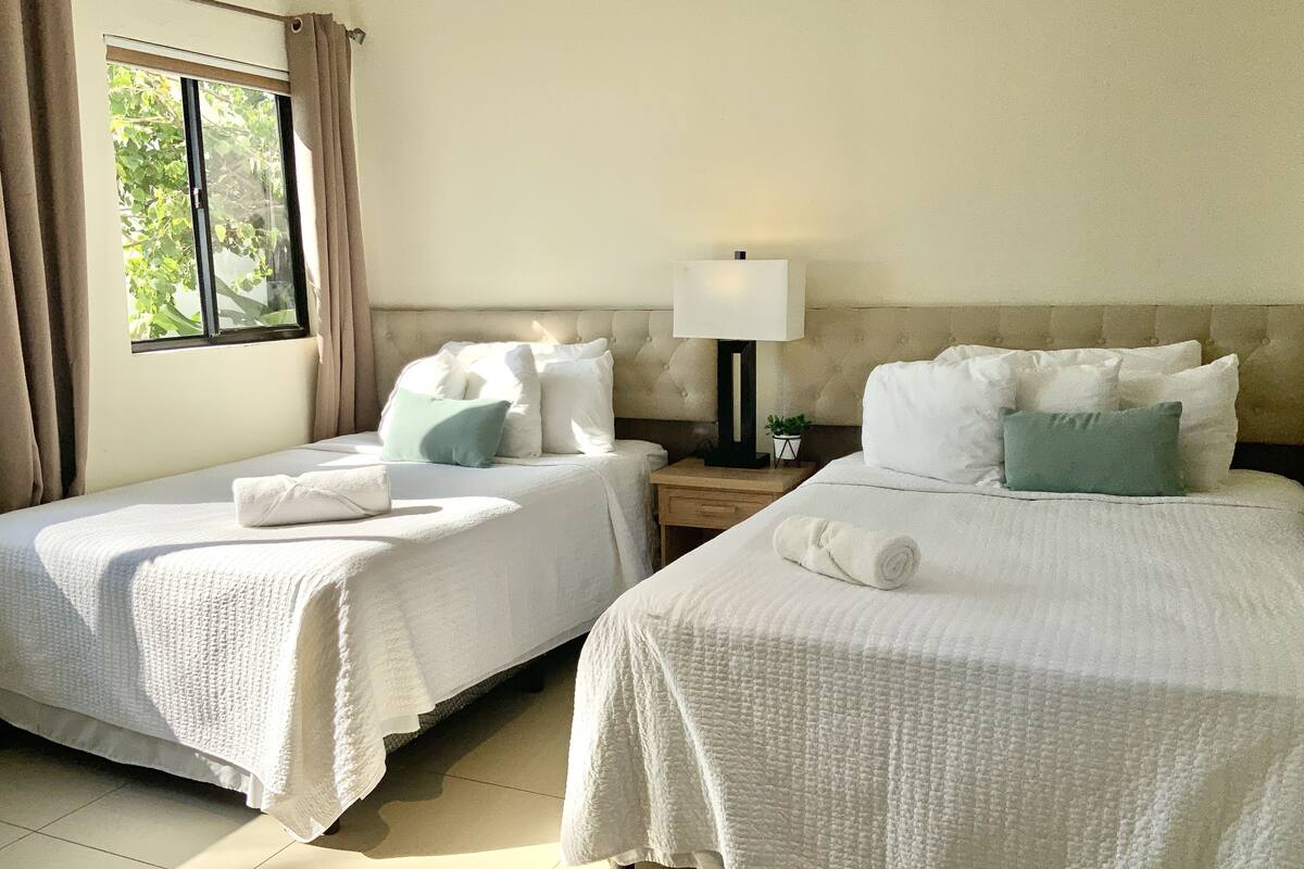 Una habitación de hotel con dos camas, una mesita de noche con lámpara y una ventana con cortinas.