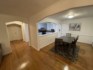 Un grand espace à manger avec une table et des chaises en bois, une cuisine à l’arrière-plan et un couloir menant à une porte.