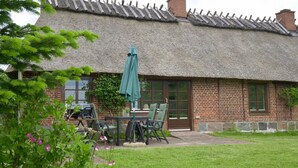 A thatched-roof building with a brick exterior, a patio with outdoor furniture, and a garden with flowering plants. - Reetdachkate im Herzen Angelns (Schnarup-Thumby)