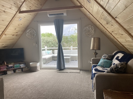 A cozy attic bedroom with a sloped ceiling, a large window with curtains, a TV, a sofa with cushions, and a lamp.
