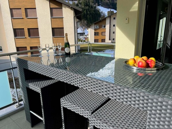A glass-top outdoor bar with seating, a bottle of champagne, and a fruit bowl.