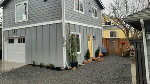 Uma casa de dois andares com fachada cinza, porta de garagem branca e pequeno jardim frontal com pedrisco, vasos de plantas e porta amarela.