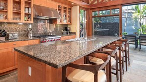 Une cuisine dotée de plans de travail en granit, de meubles en bois, d’une hotte en acier inoxydable et d’un grand îlot central avec des tabourets de bar.