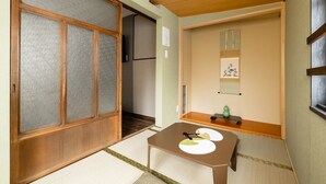 Ein traditioneller japanischer Raum mit Tatami-Boden, einer Schiebetür, einem kleinen Tisch mit Unterlage und einem an der Wand befestigten Regal mit einer Vase.