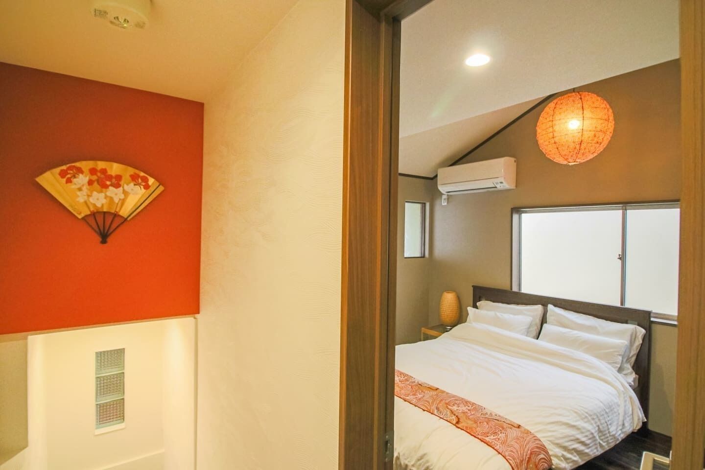 A hotel room with a bed, a wall-mounted air conditioner, a bedside lamp, and a decorative fan.