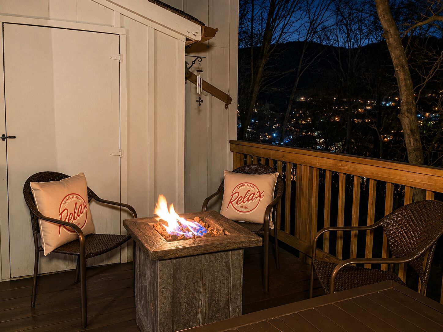 An outdoor seating area with two wicker chairs, a small table with a fire, and a view of a cityscape at night.
