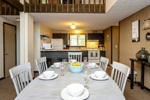 Une salle à manger avec une grande table dressée pour six personnes, entourée de chaises blanches. Un bol bleu contenant des citrons est placé au centre.