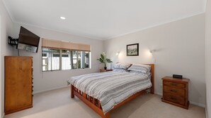 Une chambre à coucher comprenant un lit, des tables de chevet, une armoire en bois, une fenêtre avec des stores, un téléviseur fixé au mur et un tableau encadré.