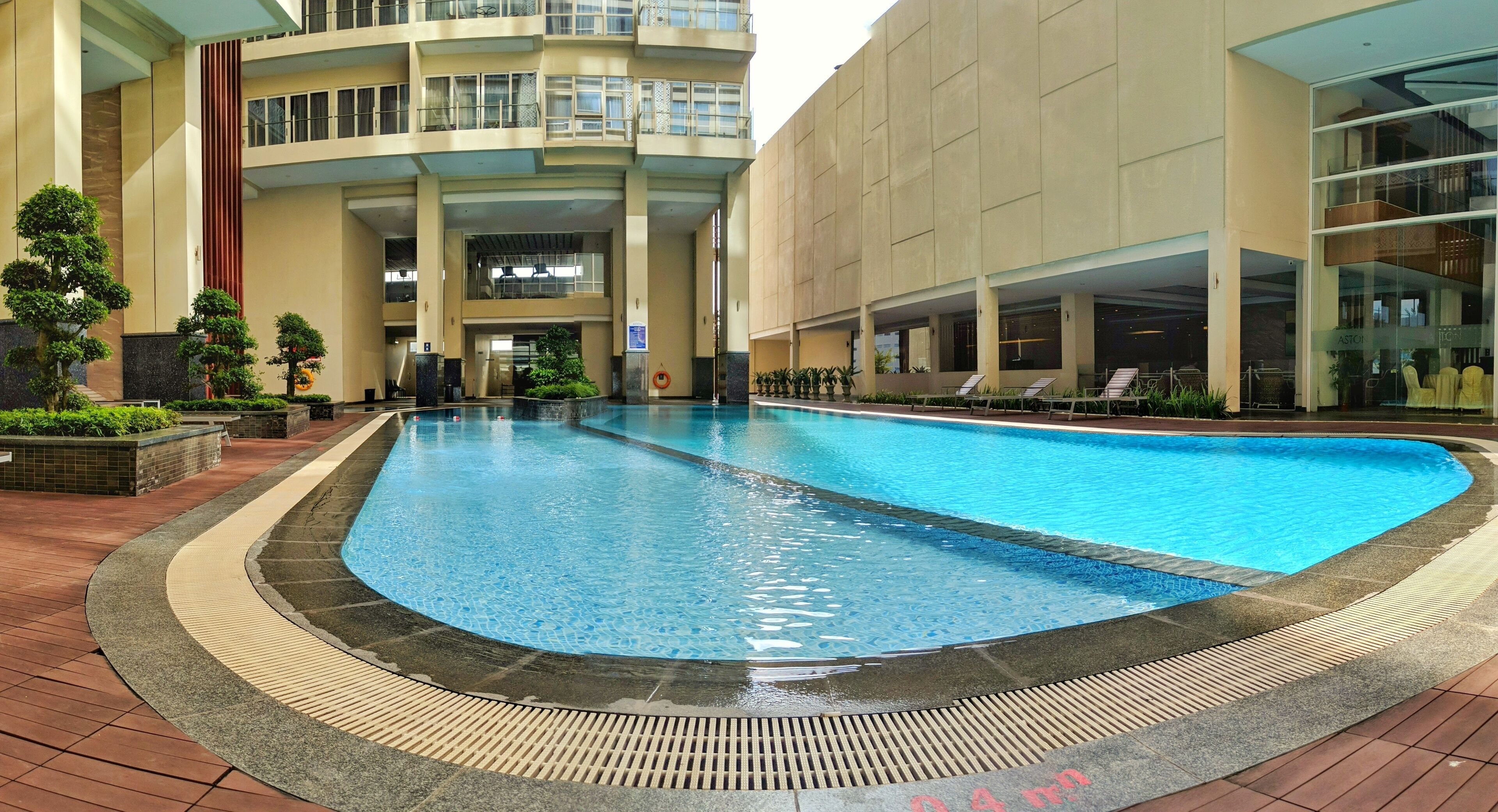 A swimming pool with a tiled deck and a building with multiple floors and balconies in the background.