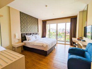 A hotel room with a large bed, a desk with a chair, a television, and a view of the city through the windows. - Lovina at Pelita Nagoya Apartment (Batam)