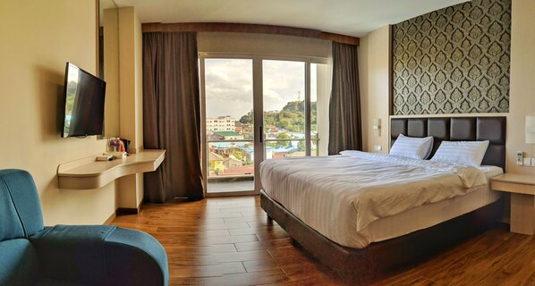 A hotel room with a bed, a TV, a desk, and a view of buildings through the window. - Lovina at Pelita Nagoya Apartment (Batam)