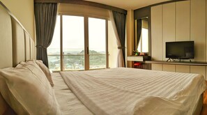 A hotel room with a large bed, a TV, a desk with a chair, and a view of the city through large windows.