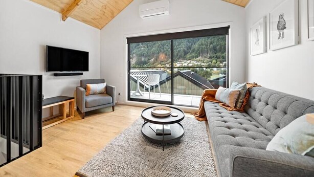 A modern living room with a grey sofa, a round coffee table, and a large window offering a view of a balcony and greenery. - 4 Bedroom, Brand New, Modern Mountain View Paradise (Queenstown)