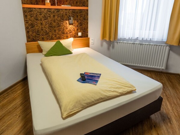 Una camera d'albergo con un letto, una testiera, una lampada a parete, una finestra con tende e un termosifone.