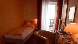 A small hotel room with a single bed, a chair, a desk with a lamp, and a window with curtains. - Einzelzimmer mit Balkon - Hotel & Ferienwohnungen Seeschlößchen (Waldeck)
