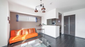 A modern living room with a glass coffee table, an orange sofa, and a kitchen area in the background. - Gravity (Sapporo)