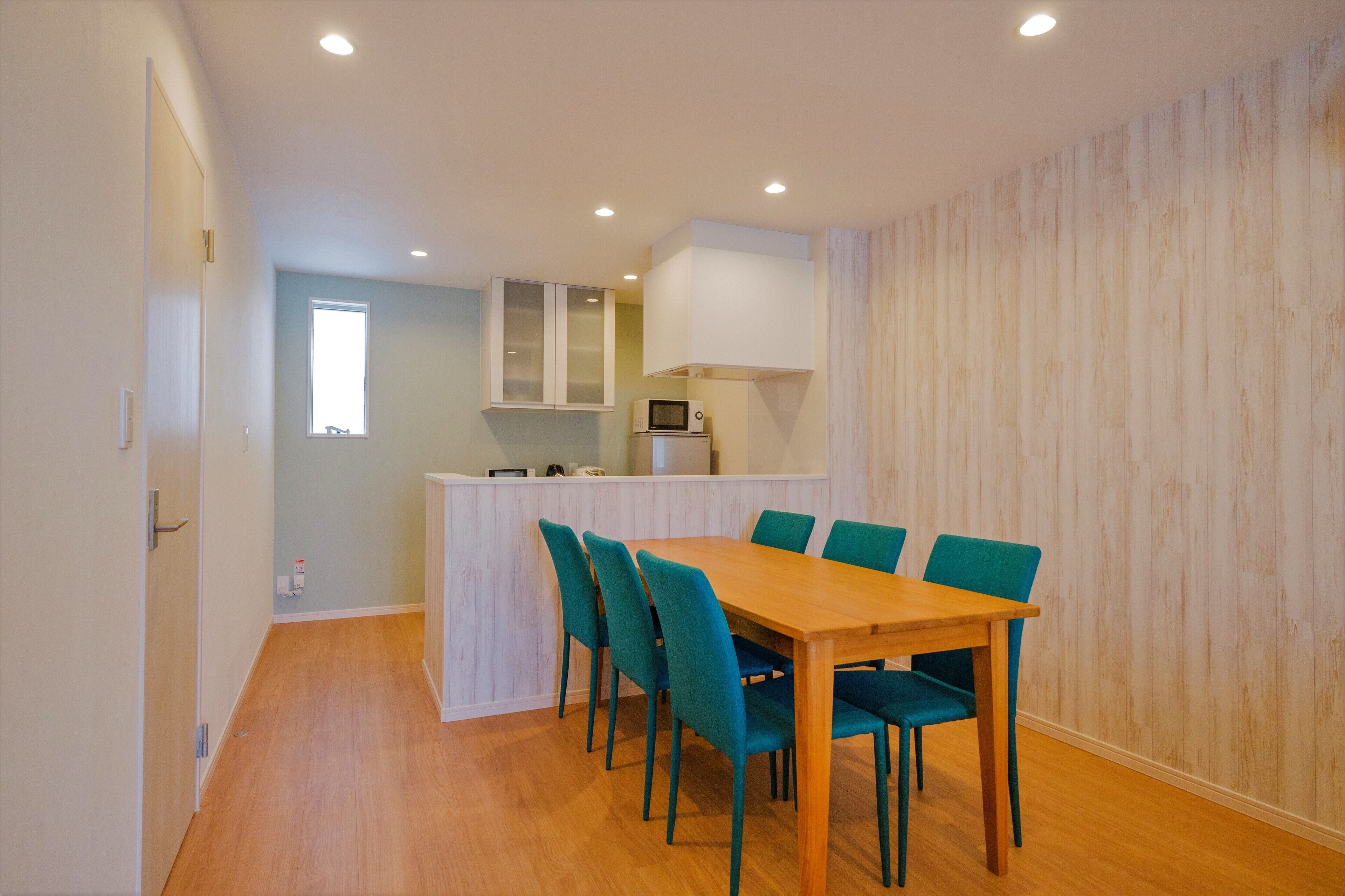 A dining area with a wooden table and teal chairs, a kitchen with a microwave, and a light wooden wall.