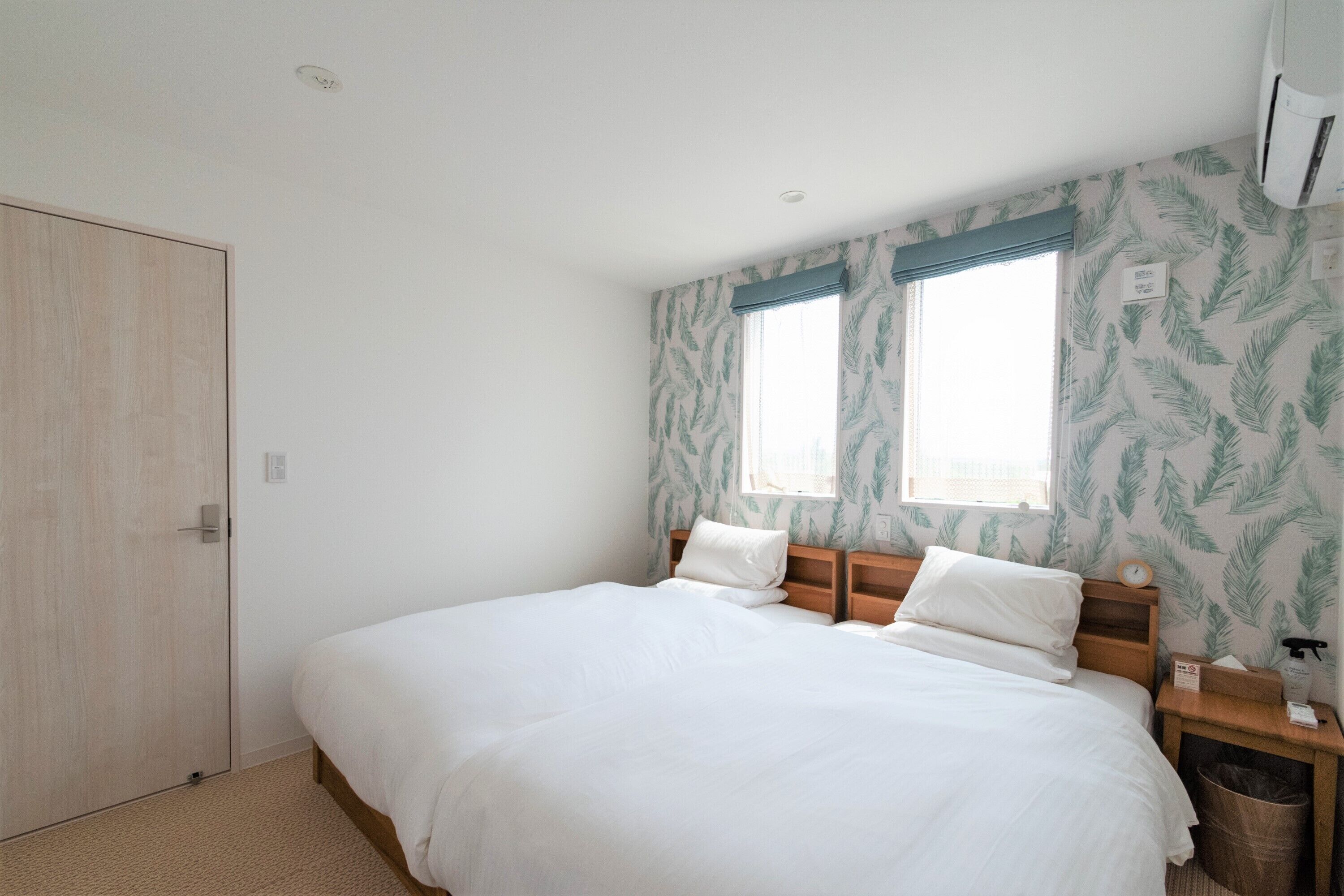 A bedroom with a large bed, a wooden wardrobe, a nightstand with a lamp, and a window with a leaf-patterned curtain.